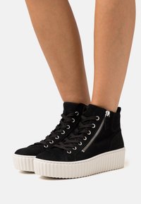 Mustat mokkanahkaiset korkeakärkiset tennarit, joissa on valkoinen platform-pohja, etulankat ja sivuvetoketju.