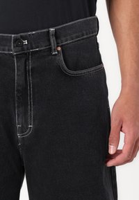 Sorte denimjeans med kontrasterende hvid syning, kobberdetaljer på lommerne og lige ben. Ingen synlig slidtage eller mønstre.