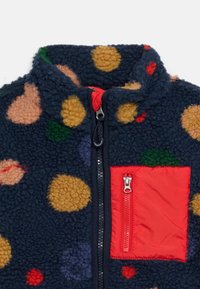 Navy fleece jacket with multicolored circular patterns, black front zipper, and red chest pocket with vertical zipper.