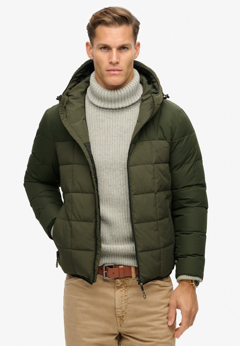 Giacca a vento verde con cappuccio, caratterizzata da un design trapuntato e dettagli neri. Indossata sopra un maglione a coste grigio chiaro e pantaloni beige.