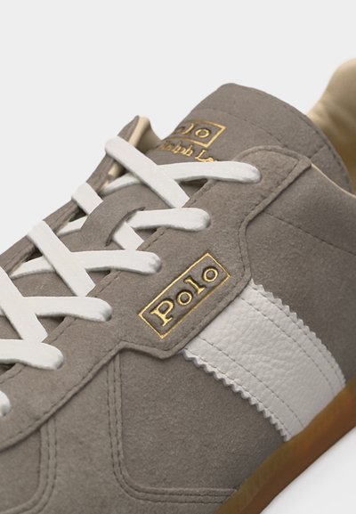 Nahaufnahme eines grauen Wildledersneakers mit weißen Schnürsenkeln, weißem Seitenstreifen und goldgeprägtem "Polo" Logo an der Seite.