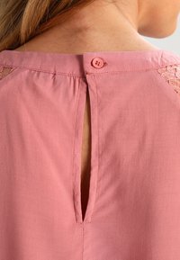 Bakifrån vy av en rosa blus med knapppunktion och spetsdetalj vid axlarna, som visar en liten nyckelhålsöppning.