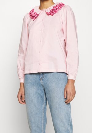 Kobieta w jasnoróżowej bluzce z długim rękawem, z haftowanym czerwonym kwiatowym kołnierzem, oraz w jasnoniebieskich dżinsach z wysokim stanem.