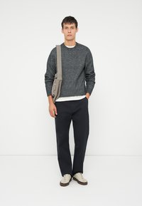 Γκρι υφασμάτινο πουλόβερ, λευκό πουκάμισο από κάτω, σε συνδυασμό με σκούρες παντελόνες. Το μοντέλο κρατά μια μπεζ τσάντα, τα παπούτσια είναι ανοιχτού χρώματος με καφέ λεπτομέρειες.