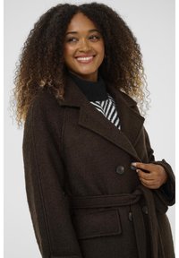 Bruine wollen blend jas met een riem in de taille, dubbelzijdige knoopsluiting, grote geknipte kraag en een gestructureerde stof. Voorzien van zwarte knopen en zakken.