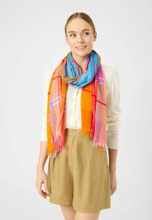 Young woman with beige shorts and white shirt wearing a bright multicolored scarf with orange, pink, blue, and green tones.