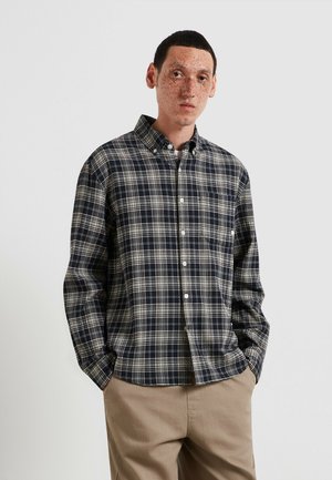 Giovane con capelli corti e scuri e lentiggini, che indossa una camicia a quadri nera, grigia e bianca con bottoni e pantaloni beige, con le mani nelle tasche.