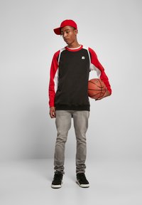Felpa a maniche lunghe nera e rossa con dettagli bianchi, jeans grigi e sneakers nere. Tiene in mano un pallone da basket, indossando un berretto rosso inclinato di lato.