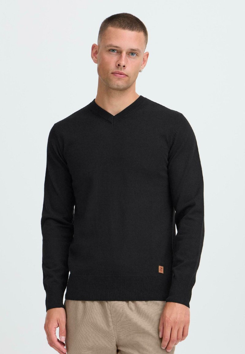 Man met kort haar in een zwarte V-hals trui en beige broek, tegen een effen lichte achtergrond, recht vooruit kijkend.