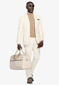 Hvid fløjlssæt med en struktureret beige sweater, brune handsker og bærende en lys beige duffelbag med læderdetaljer.