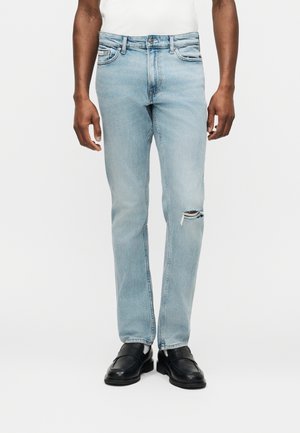 Mężczyzna ubrany w jasnoniebieskie, dopasowane jeansy z małym rozdarciem na prawym kolanie, w połączeniu z czarnymi skórzanymi mokasynami i białą koszulą.