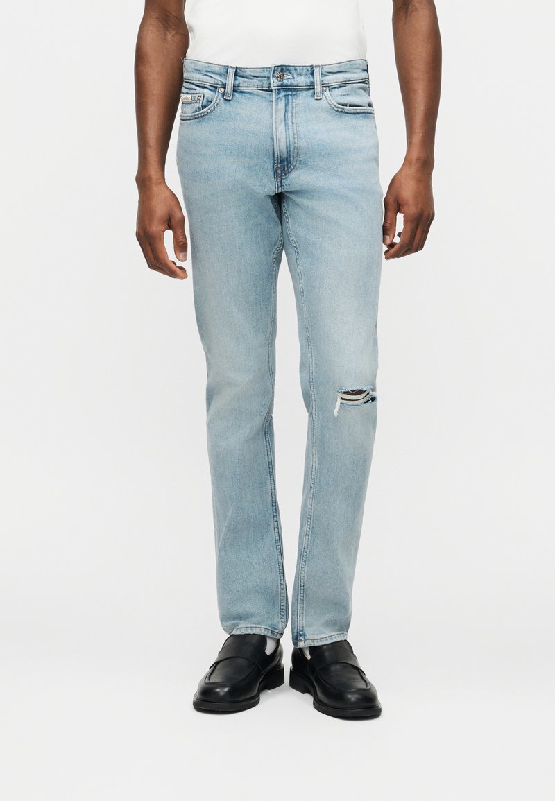 Άνδρας που φοράει ανοιχτό μπλε τζιν σε στενή εφαρμογή με μικρό σκίσιμο στο δεξί γόνατο, συνδυασμένο με μαύρα δερμάτινα loafers και λευκό πουκάμισο.