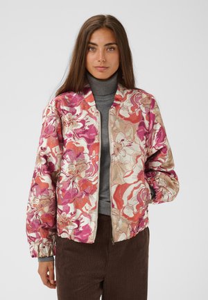Frau mit langen braunen Haaren, die eine florale rosa, rote und beige Jacke über einem grauen Rollkragenpullover und braunen Hosen trägt, steht vor einem schlichten Hintergrund.