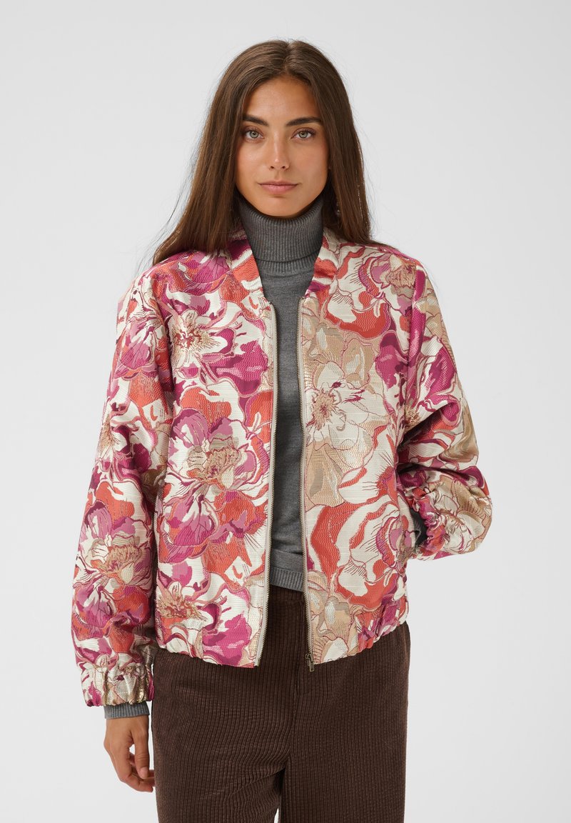 Femme aux longs cheveux bruns portant une veste imprimée florale rose, rouge et beige par-dessus un col roulé gris et un pantalon marron, debout devant un fond uni.