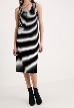 Vrouw die een mouwloze jurk draagt met zwart-witte horizontale strepen tot op de knie, gecombineerd met zwarte enkellaarzen.