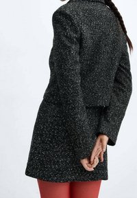 Blazer noir texturé avec un design court, motif moucheté et manches longues. Porté sur une jupe ajustée, les mains croisées dans le dos.