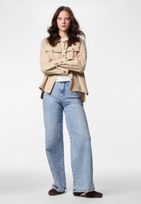 Beigen värinen ribbineulottu paitatakki, jossa läppätaskut, valkoinen t-paita alla, vaaleansiniset leveälahkeiset farkut ja tummat liukusukat.