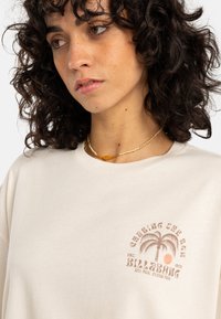Lys beige bomull T-skjorte med rund hals, med et palmetre-grafikk og "BILLABONG"-tekst i brun. Korte ermer og avslappet passform.