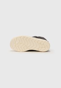 Zapatilla negra con parte superior texturizada y suela de goma blanca con patrones de zigzag. Incluye detalles como el logotipo en el lateral.