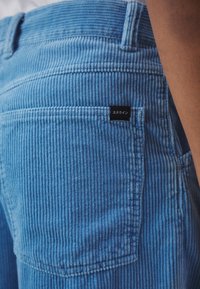 Gros plan d'une personne portant un pantalon en velours côtelé bleu clair avec une poche arrière avec une petite étiquette noire portant un texte japonais.