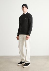 Melnas adītas svītras, zem kurām ir balta krekla, off-white bikses, melni sporta apavi ar biezu zoli un melna beanie cepure. Neitrāla fona.