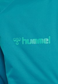 Türkisfarbener Stoff mit einem kleinen Bienenlogo und dem Wort "hummel" in hellgrün auf texturiertem Sportmaterial gedruckt.