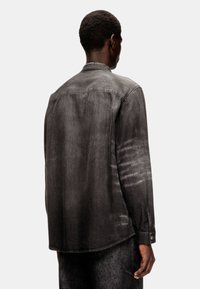 Chemise en denim noir à manches longues, légèrement brillante, avec des poignets boutonnés et un ourlet arrondi, présentant une texture délavée et un design minimaliste.