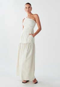 Jasny beżowy, prążkowany top na jedno ramię w połączeniu z luźną, warstwową spódnicą maxi. Posiada kieszenie i szeroki pas. Płaskie czarne sandały.