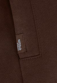 Bruin gebreide stof met een klein label waarop "spitbulb studios" staat in witte letters op een gestikte rechthoekige patch. Zachte textuur, eenvoudig ontwerp.