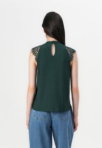 El modelo lleva una camiseta sin mangas de color verde oscuro con detalles de encaje en los hombros, combinada con unos vaqueros holgados azules, vista desde atrás.