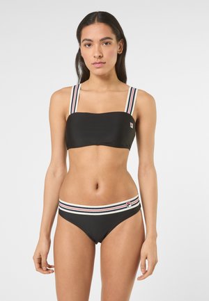 Donna che indossa un costume da bagno nero a due pezzi con spalline e fascia in vita a righe, in piedi davanti a uno sfondo chiaro uniforme, rivolta verso la fotocamera.