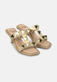 Sandali con tacco beige con cinturini incrociati, adornati con borchie piramidali in metallo oro e argento, con punta quadrata e tacco basso a blocco.