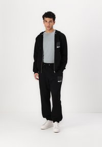 Sort zip-up hoodie med en frontal lomme, parret med sorte sweatpants med elastiske manchetter; lys grå t-shirt under, hvide sneakers.