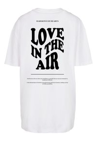 Witte T-shirt met korte mouwen met opvallende zwarte tekst die "LOVE IN THE AIR" leest, met daarboven de frase "HARMONY OF HEARTS".