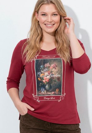 Uśmiechnięta kobieta z długimi blond włosami, ubrana w czerwoną bluzkę z długim rękawem z nadrukiem kwiatowego wazonu i tekstem "Von Landen."