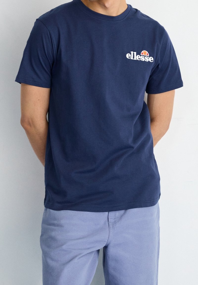 Námořnická modrá bavlněná tričko s krátkými rukávy, kulatým výstřihem a bílým logem "ellesse" na hrudi, které obsahuje červený a oranžový polokruhový design.