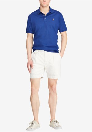 Homme portant un polo bleu, un short blanc et des baskets blanches, debout avec une main dans la poche.