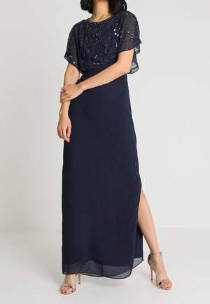 Woman wearing a navy blue floor-length dress with sheer sequined short sleeves and a side slit, paired with nude high-heeled sandals.