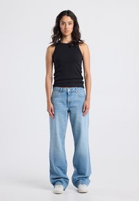 Young woman with curly dark hair wearing a black sleeveless top, light blue baggy jeans, and white sneakers standing against a plain background.