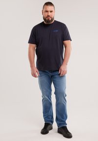 Habemega mees seisab, kannab musta "Big Star" T-särki, helesiniseid teksapükse ja musti tenniseid, taustal on hele ühtlane sein.