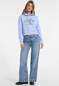 Felpa corta azzurro chiaro con logo e scritta bianca, abbinata a jeans azzurro chiaro a zampa d'elefante. Scarpe nere completano l'outfit.