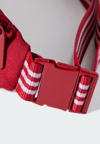 Červený nastavitelný popruh s plastovou sponou, zdobený bílými a červenými pruhy a s texturovaným povrchem pro lepší úchop.