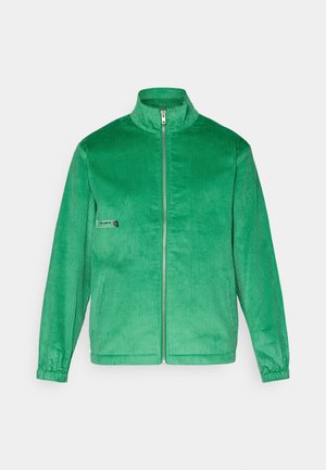 Zielona kurtka w sztruks z wysokim kołnierzem, pełnym zamkiem z przodu, elastycznymi mankietami i kieszenią naszywaną na lewej piersi. Teksturowana powierzchnia.