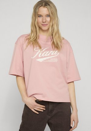 Młoda kobieta o długich blond włosach, ubrana w różowy oversize T-shirt z białym napisem oraz ciemnobrązowe spodnie, stojąca na jednolitym tle.