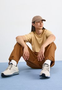 Beige Nike póló a logójával, barna szűkített nadrág, bézs és fekete sportcipő, fehér zokni, és könnyű barna sapka. Kék felületen ül.