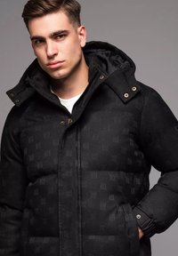 Svart puffjacka med ett mönstrat textur, dragkedjestängning och vikbar huva. Har sidofickor och guldsmycken på knapparna.