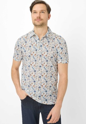 Mand med lyst brunt hår iført en kortærmet hvid skjorte med blå og brune blomsterprint og mørkeblå jeans, stående mod en hvid baggrund.