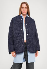 Navy quilted jacket with a textured pattern, featuring snap buttons, a round collar, and a relaxed fit, worn over a light blue shirt.