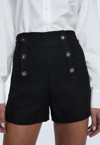 Černé tweedové šortky s texturovaným povrchem, vysokým pasem a výrazným předním detailem se dvěma knoflíky. V kombinaci s bílou košilí s límečkem.