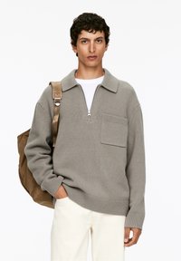 Grauer Strickpullover mit Reißverschlusskragen, kurzem Kragen und Seitentasche. Über einem weißen Hemd getragen, kombiniert mit cremefarbenen Hosen und einem braunen Rucksack.